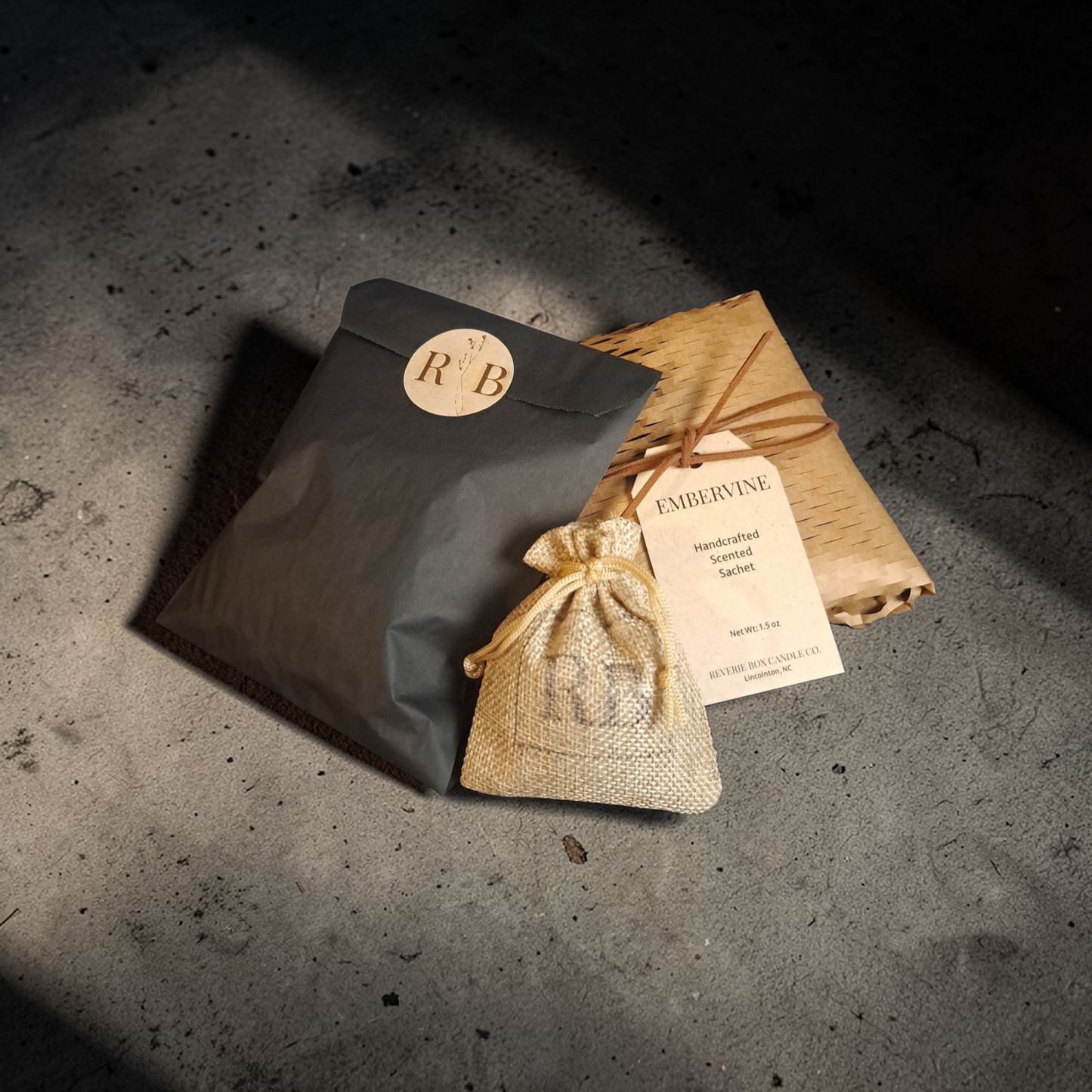 Dark charcoal package with RB branding, smooth beige drawstring RB branded burlap bag, and kraft honeycomb paper on a textured concrete surface with dramatic shadows.