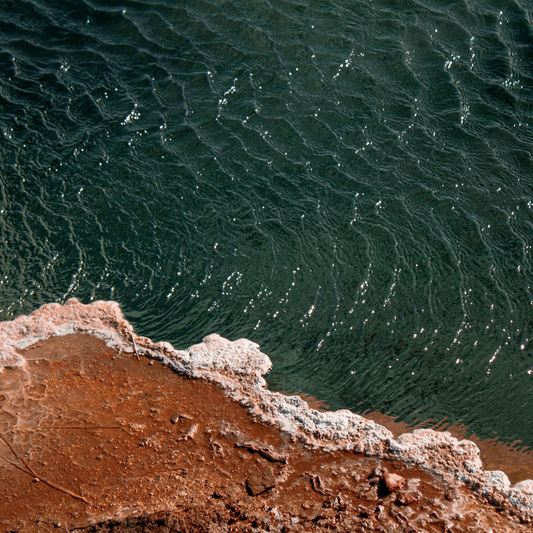 A moody, high-contrast overhead shot of a rough, white sea salt mineral crust along a textured shoreline ledge cutting against deep, dark shimmering azure ocean water.