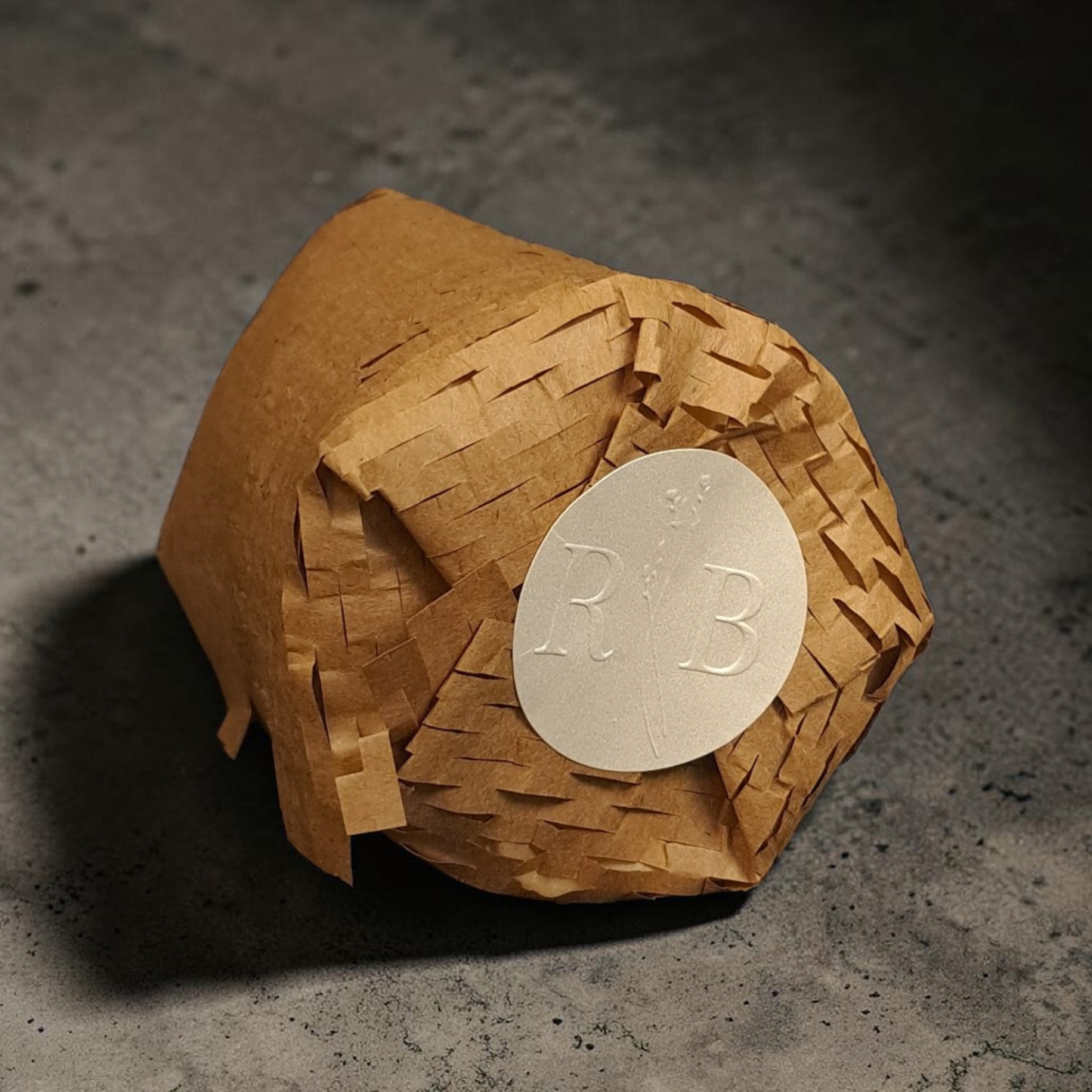 Candle refill insert wrapped inside of kraft brown honeycomb paper with a RB branded pearlized sticker label lying sideways on a concrete surface.