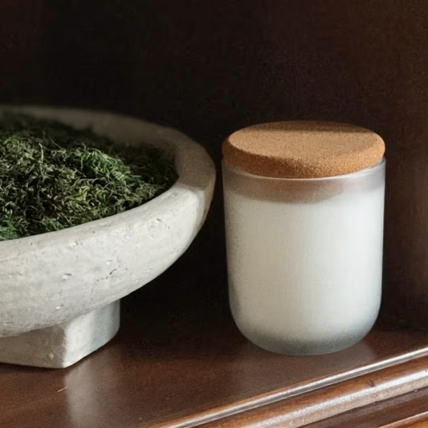 A frosted white glass Reverie Box Orriswood candle jar with a natural cork lid, sitting on a dark wooden shelf next to a concrete bowl of green moss in minimalistic home.
