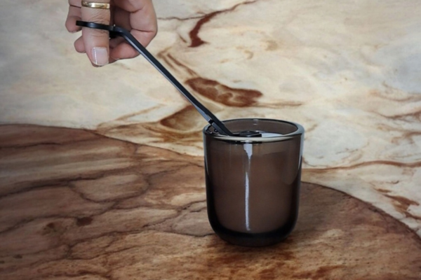 A hand trimming the wicks of a deep brown Reverie Box Mossfall glass candle vessel on a mocha and cream marbled setting.