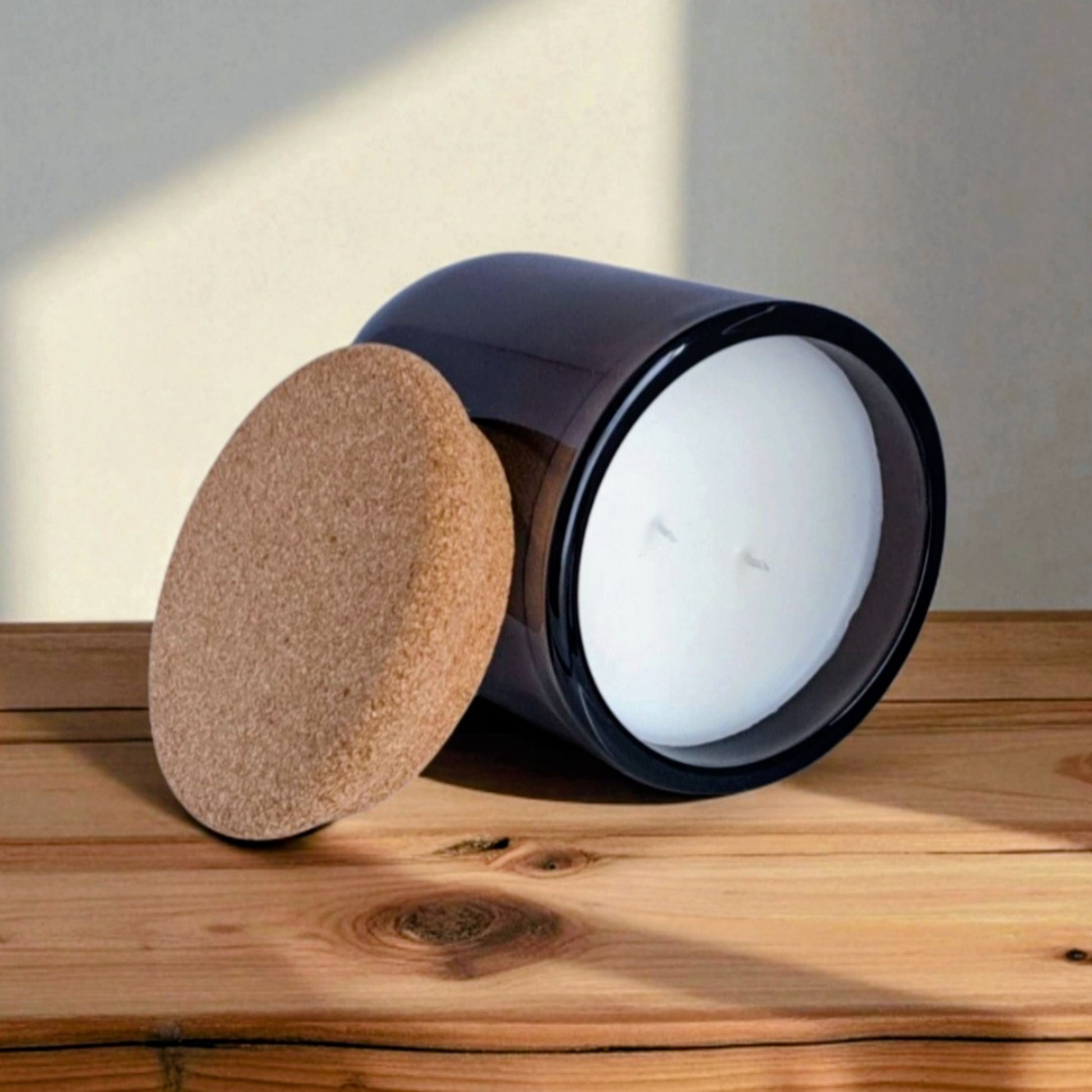 A dark brown, unlit Reverie Box Mossfall glass candle with cork lid, placed on a wooden ledge with warm sunlight and shadows.
