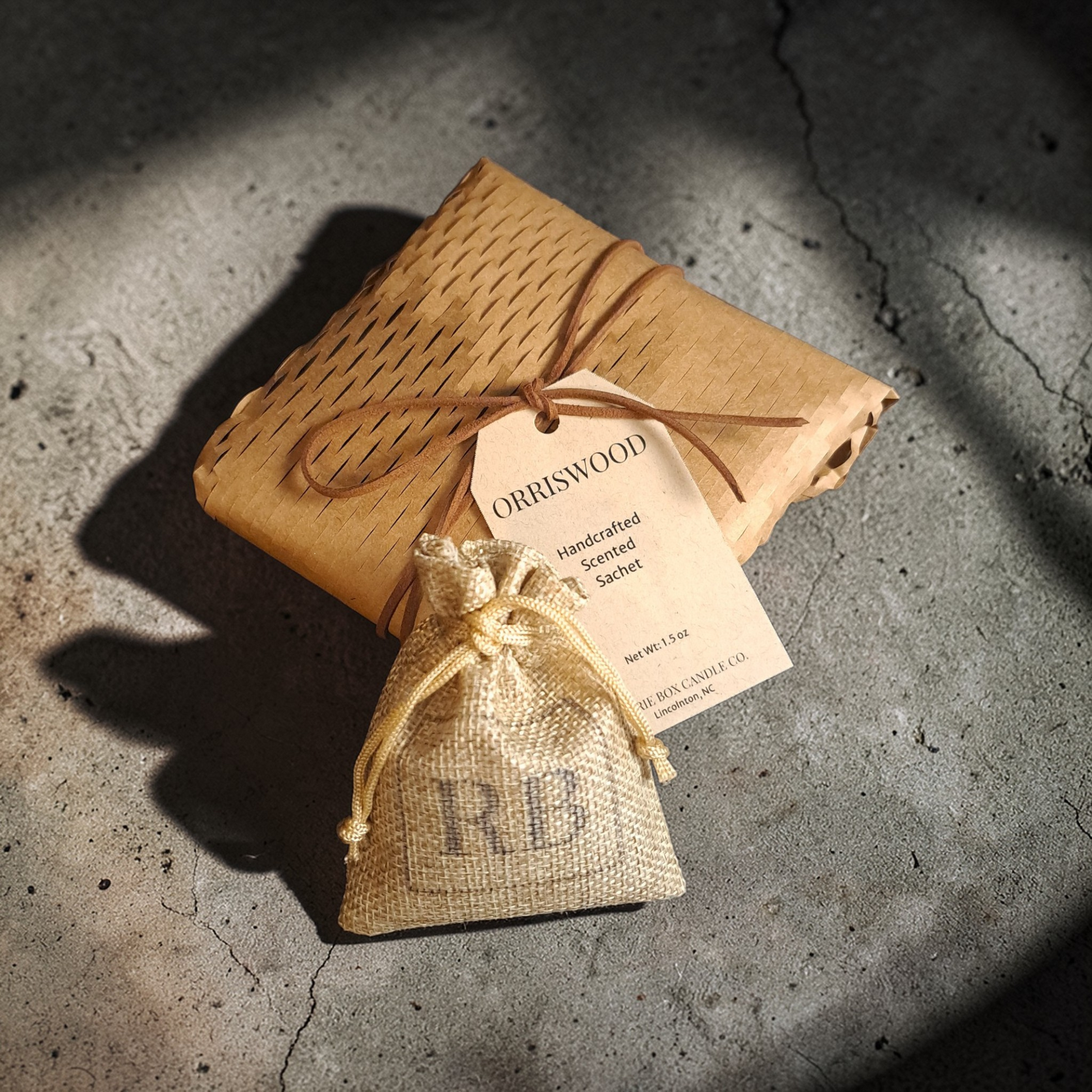 Handcrafted scent sachet in a soft RB branded burlap bag with a kraft label on a textured concrete surface with dramatic light and shadows.