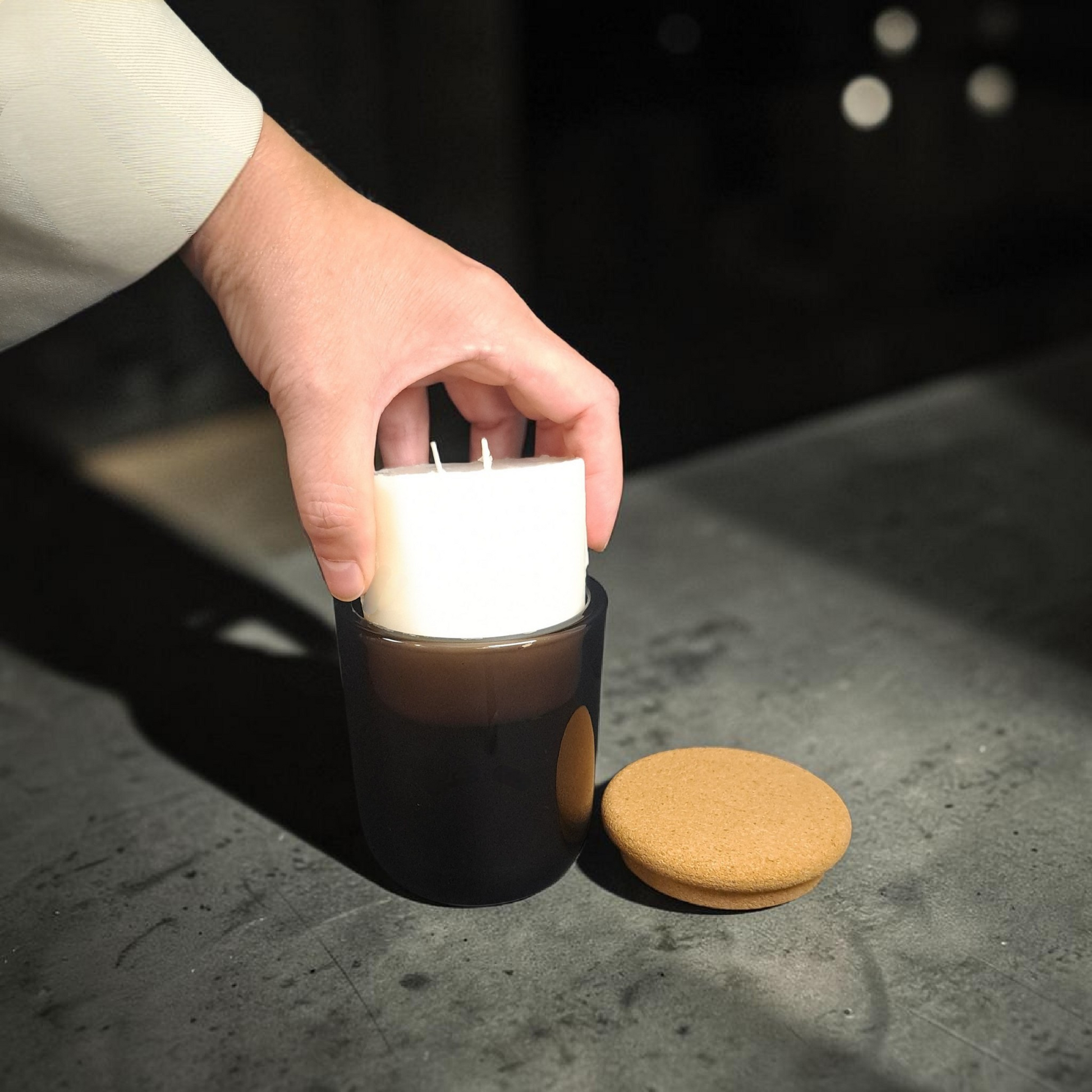 Hand holding and easing coconut apricot wax refill insert down into dark brown glass vessel next to cork lid on concrete surface.