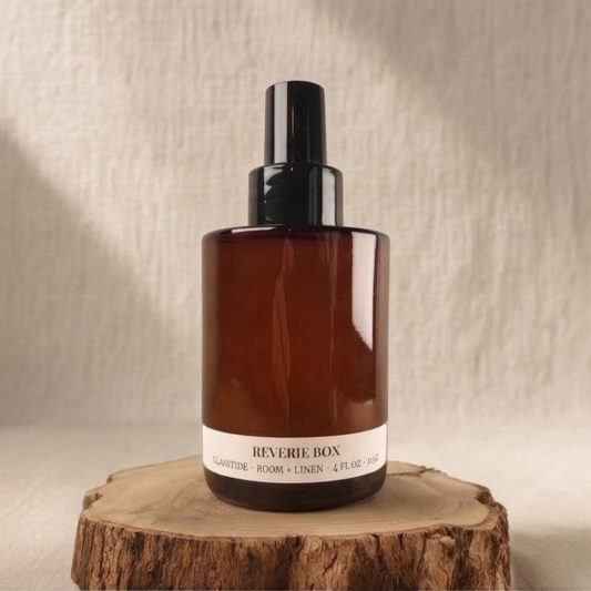 Amber glass spray bottle with black cap labeled  "REVERIE BOX GLASSTIDE ROOM + LINEN 4 FL OZ 113G", sitting on a natural wood slice with rough bark edges against a soft beige fabric background.