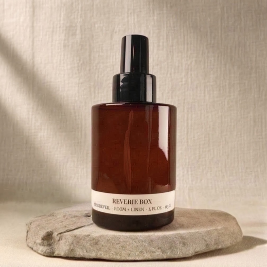 Amber glass spray bottle with black cap labeled  "REVERIE BOX SHOREVEIL ROOM + LINEN 4 FL OZ 113G", sitting on a flat gray stone base against a soft beige fabric background.