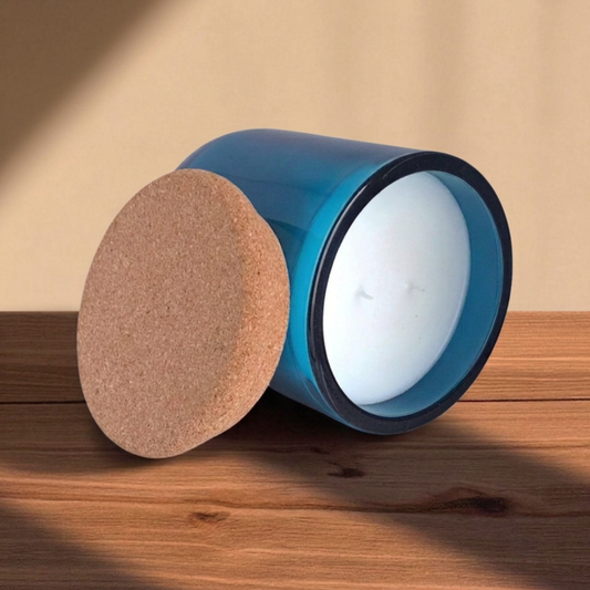 An unlit Reverie Box Saltmere blue glass candle with a round cork lid leaning against it detailing smooth wax top and dual wicks.