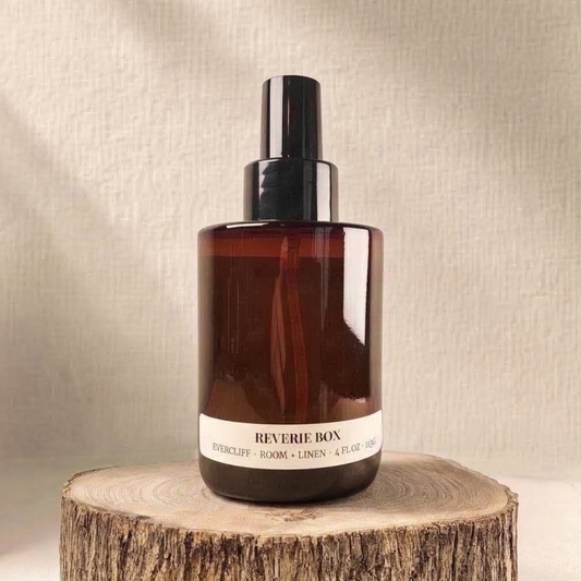 Amber glass spray bottle with black cap labeled "REVERIE BOX EVERCLIFF ROOM + LINEN 4 FL OZ 113G", sitting on a natural wood slice with bark edges against a soft beige fabric background.
