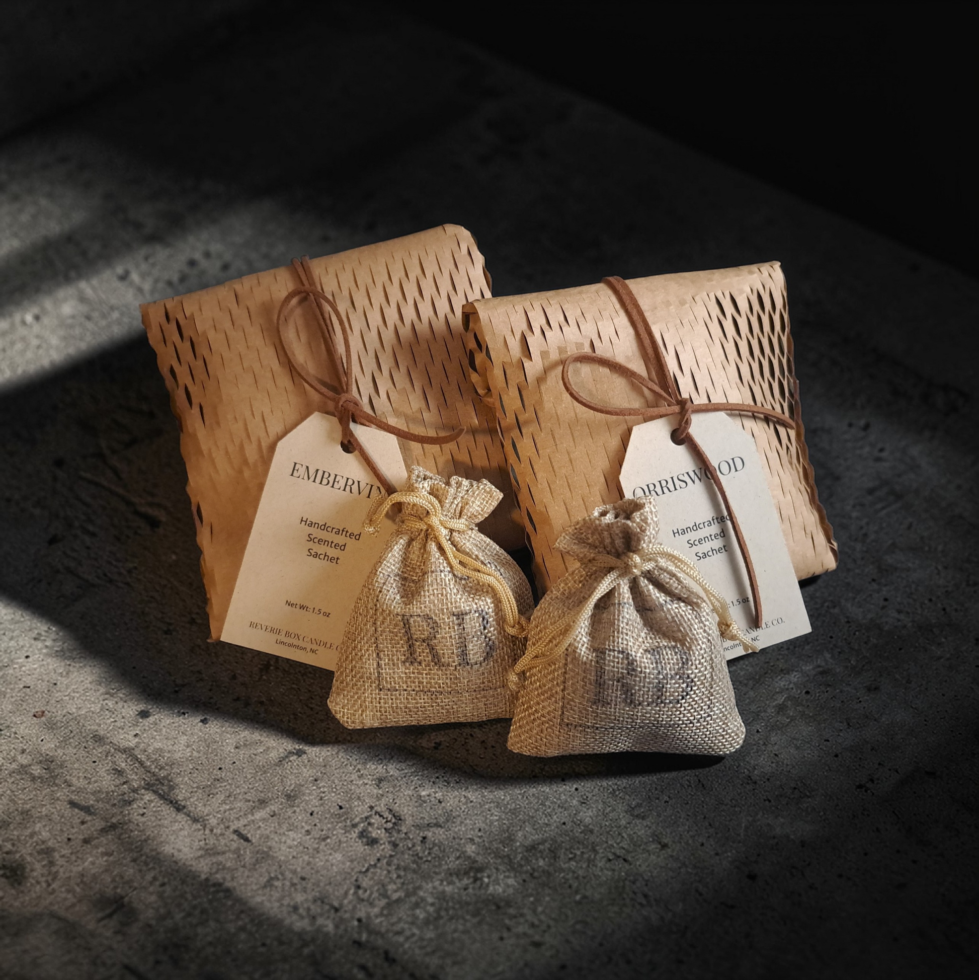 Two honeycomb wrapped Reverie Box scent sachets with drawstrings and kraft tags and leather cords on a concrete surface