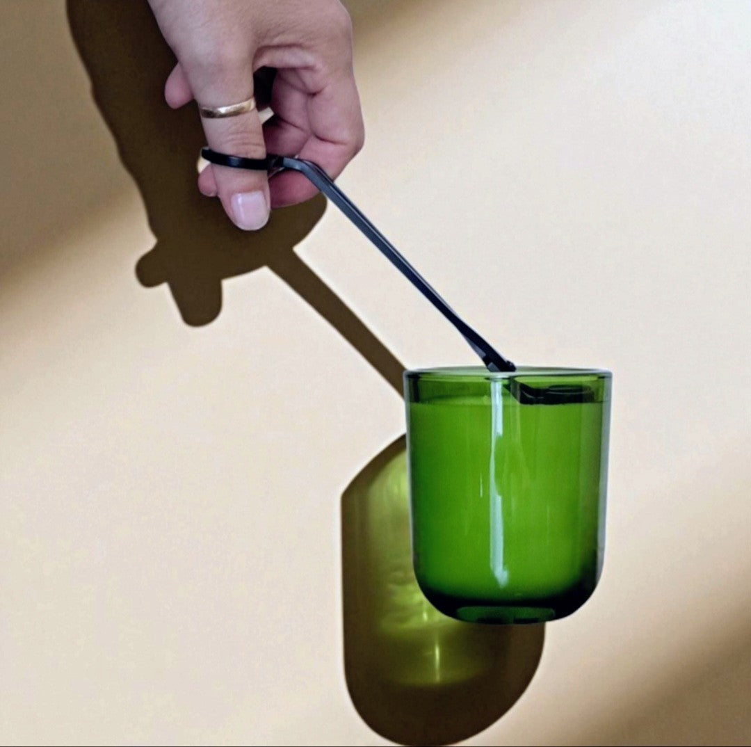 Hand trimming the wicks of a Reverie Box Fogborn candle in vibrant green glass tumbler, casting geometric shadows against warm background.