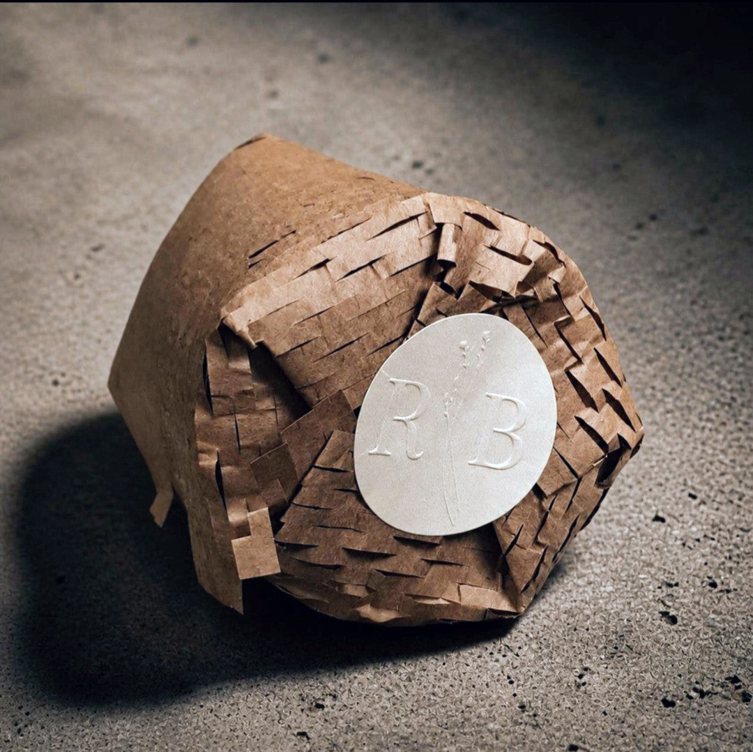 A luxury coconut-apricot wax candle refill insert wrapped in sustainable brown honeycomb packaging paper, sealed with a round, RB embossed pearlized label lying on a dark concrete background.