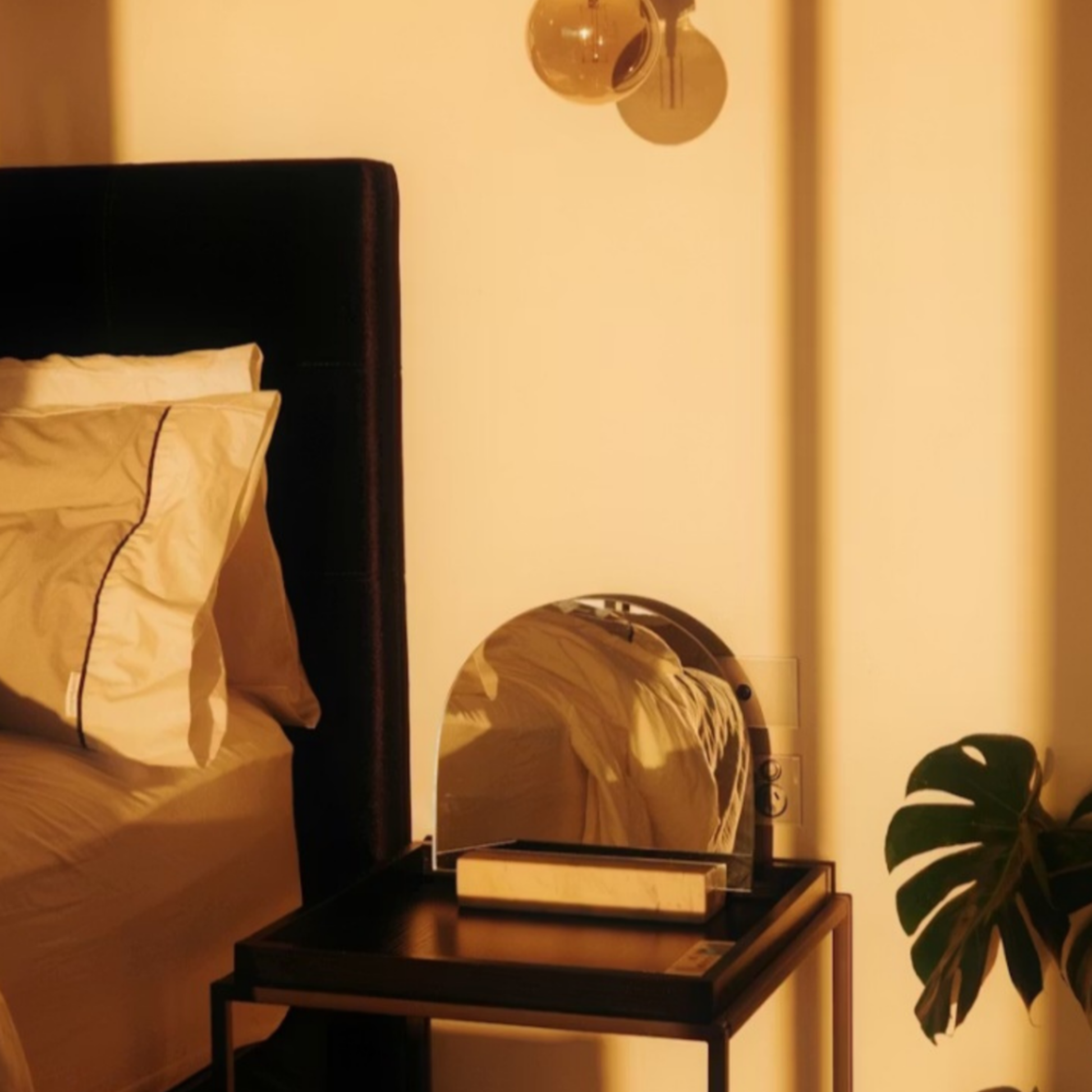 Warm golden-lit bedroom with black upholstered bed, arched mirror on wooden nightstand, monstera leaf, and soft ambient lighting creating a romantic glow